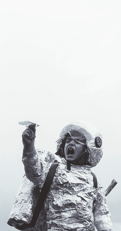 A child in a homemade foil spacesuit holding up a paper plane against an overcast sky